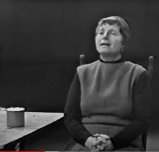 Ruth Rubin auf einem einfachen Stuhl, Blechtasse neben sich, singt.